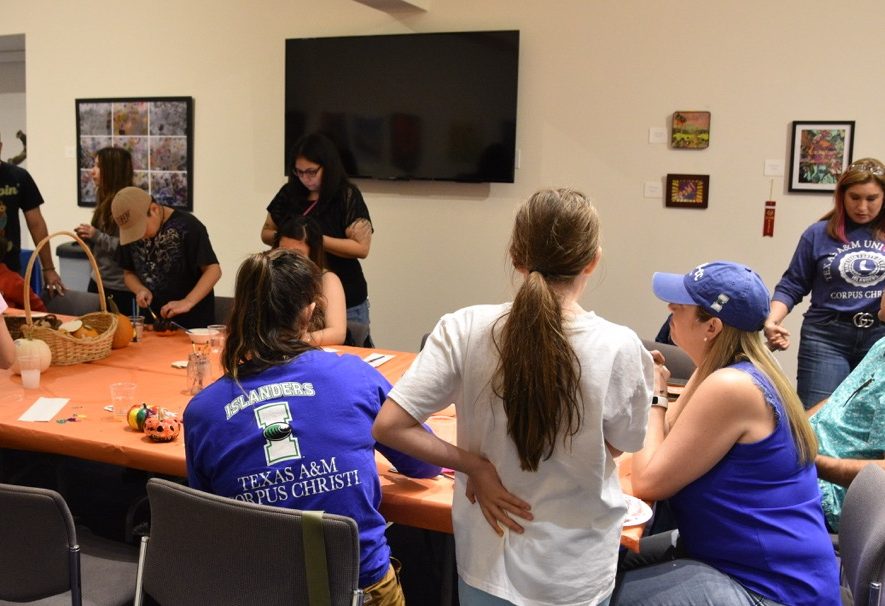 Group of Islander alumni, families, and children engaged in various activities around tables in a community center room at the Art Museum of South Texas with decorations and an Islanders Texas A&M University–Corpus Christi shirts and colors and cap are visible.
