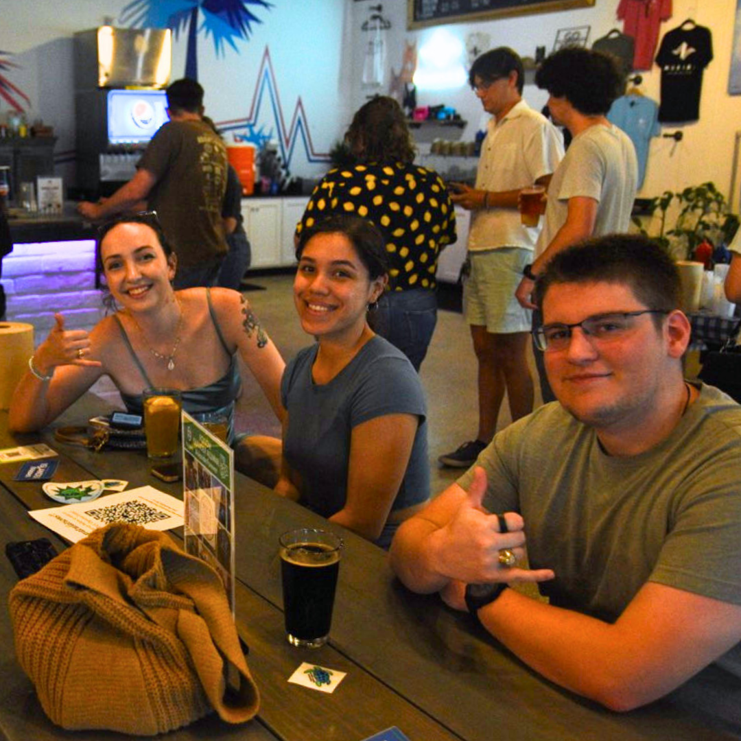 Three young Islander alumni friends smiling at a table in a casual bar setting, enjoying drinks together at the TAMU-CC Islander Alumni Association Senior Party.