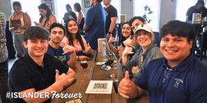 A group of young Texas A&M University-Corpus Christi graduates sitting around a table at an Islander Alumni Association gathering, smiling and giving shakas-up gestures. There is a drink on the table along with a QR code. The foreground features the text "#ISLANDERForever.