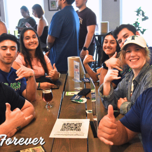 A group of young Texas A&M University-Corpus Christi graduates sitting around a table at an Islander Alumni Association gathering, smiling and giving shakas-up gestures. There is a drink on the table along with a QR code. The foreground features the text "#ISLANDERForever.