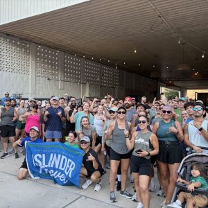 A large group of run club runners happily gather under a covered area, some posing with a "Islander Forever, Texas A&M University-Corpus Christi" banner. Several are wearing athletic gear, and sunglasses. A stroller with a child is visible in the foreground.