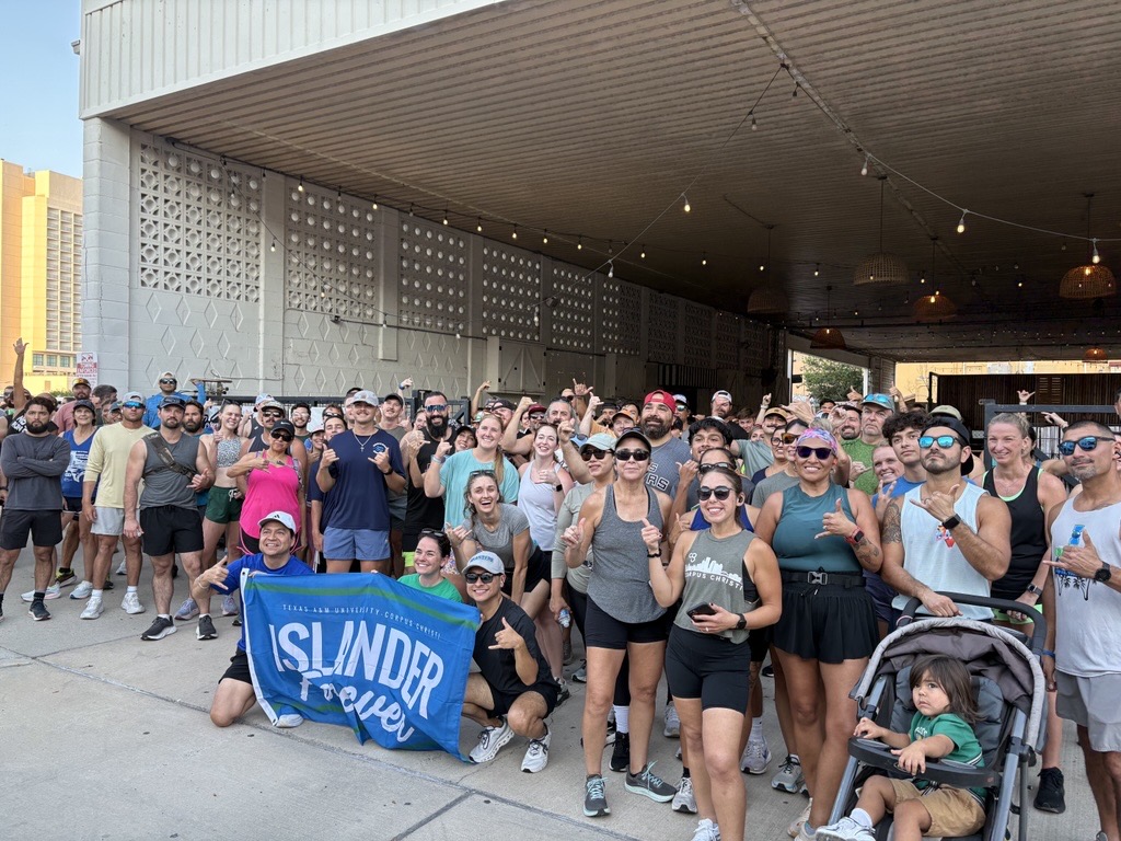 A large group of run club runners happily gather under a covered area, some posing with a "Islander Forever, Texas A&M University-Corpus Christi" banner. Several are wearing athletic gear, and sunglasses. A stroller with a child is visible in the foreground.